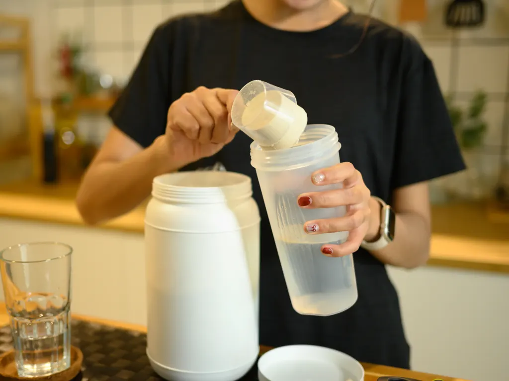 A scoop of protein powder tipped into a shaker bottle on a kitchen counter