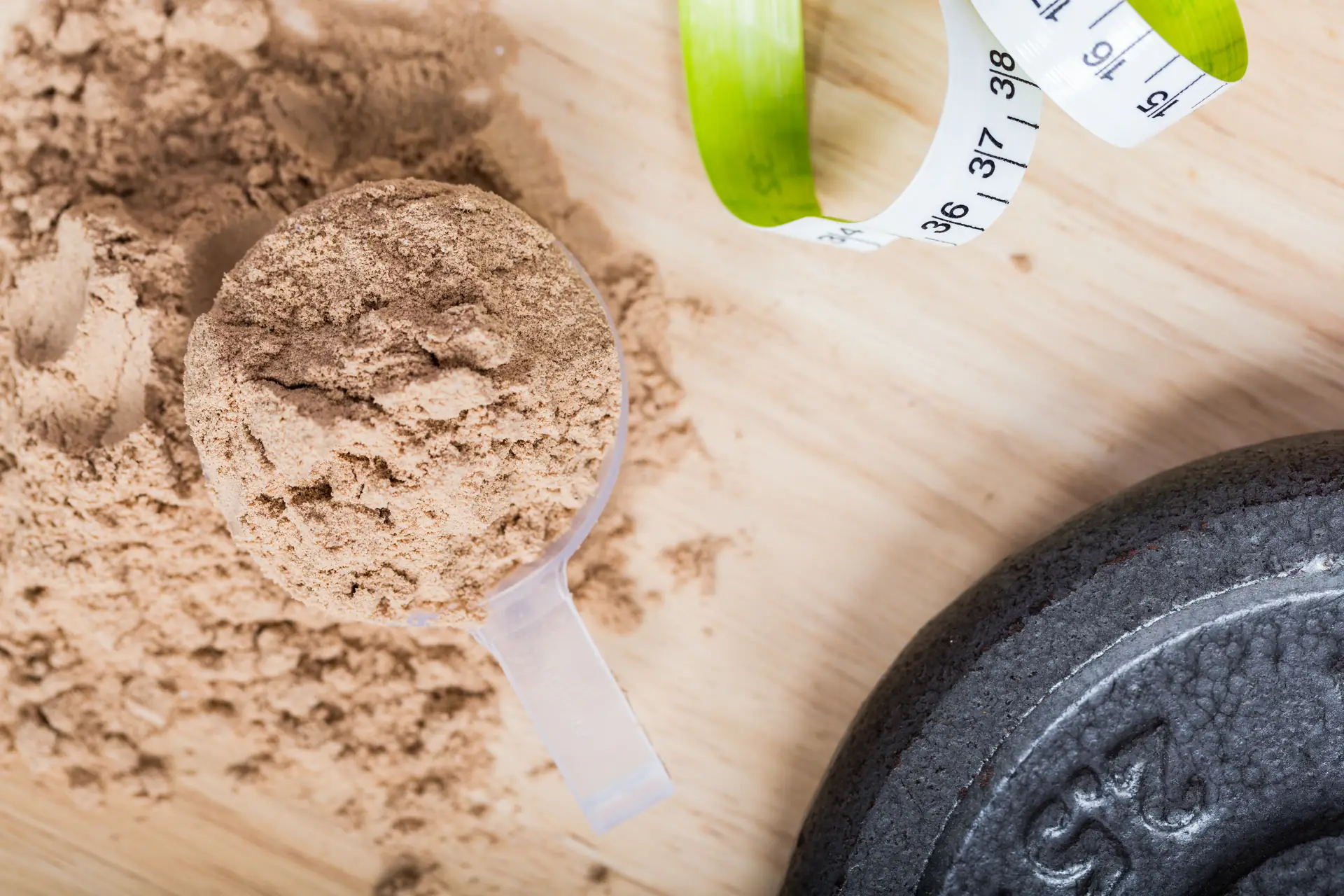 A scoop of protein powder on a wooden bench, beside a tape measure and a weight plate