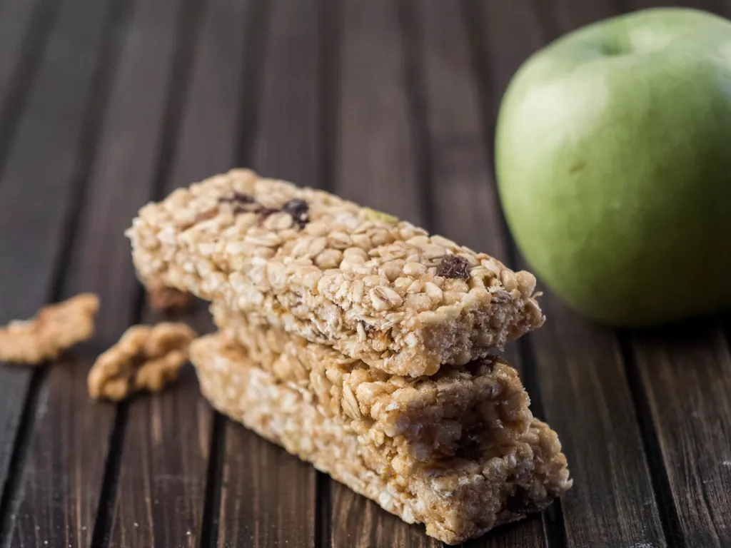 Two protein bars stacked on a wooden board with walnuts and a green apple